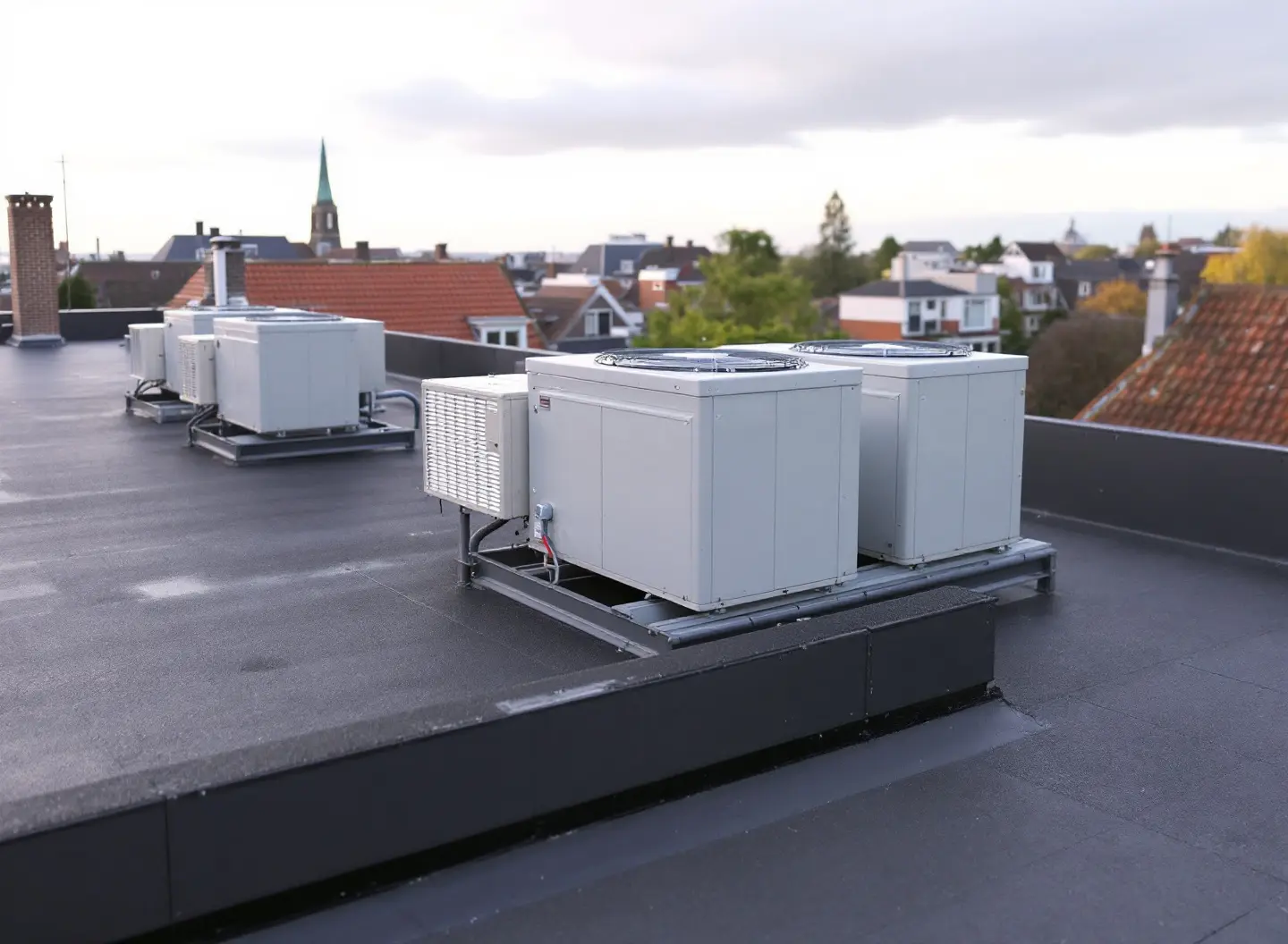 Buitenunits van warmtepompen op een stalen frame op het platte dak van een appartementengebouw, met daken en een kerktoren op de achtergrond